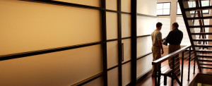 Two men looking at design documents in a modern office near a stairway and conference room